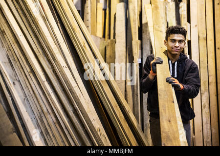 Jeune homme portant des gants de travail debout à côté d'une pile de planches de bois dans un entrepôt, l'exécution de longs morceaux de bois, en regardant la caméra. Banque D'Images