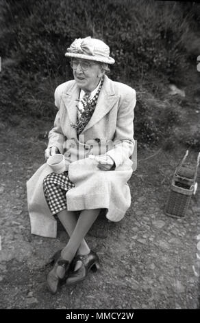 Années 50, une vieille dame bien habillé portant un chapeau et enduire assis sur un chemin à l'extérieur, avoir une tasse de thé et une collation, England, UK. Banque D'Images