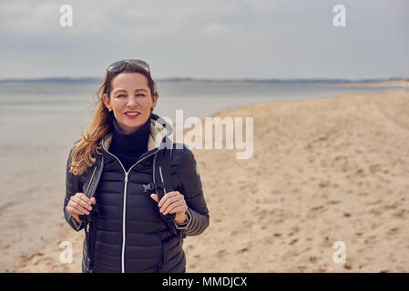 Jolie femme en bonne santé bénéficiant d'une randonnée sur la plage un jour nuageux froid debout souriant à la caméra sur le sable tenant les sangles de son retour Banque D'Images