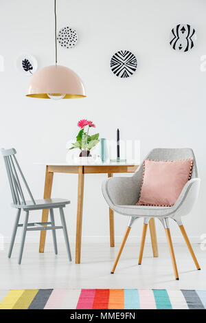 Vue de face d'une table à manger, chaise, fauteuil rétro avec un oreiller, tapis aux couleurs vives, de la lampe et les plaques sur le mur blanc Banque D'Images