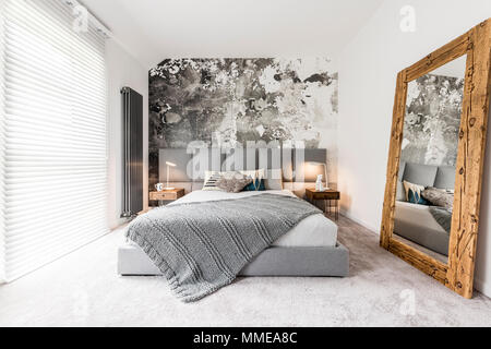 Lit king-size avec tête de lit carré gris, grand miroir en bois rustique et mur texturé dans un appartement minimaliste Banque D'Images