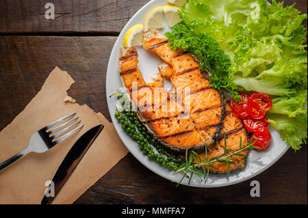 Pavé de saumon grillé servi avec des tomates grillées, salade, tranches de citron, graines de poivre, romarin, persil sur plaque blanche sur table en bois. Banque D'Images
