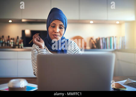 Souriante jeune femme entrepreneur musulmane portant un hijab assis à une table dans sa cuisine écrit des notes et de travailler sur un ordinateur portable Banque D'Images