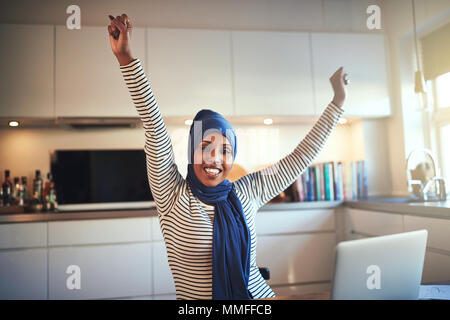 Excitée jeune musulman femme entrepreneur portant un hijab raising her arms à succès tout en travaillant dans sa cuisine sur un ordinateur portable Banque D'Images