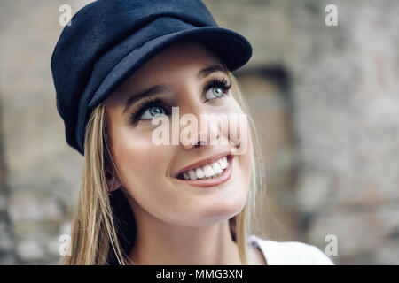 Jeune femme blonde en fond urbain de flou artistique. Fille aux yeux bleus portant le chapeau. Banque D'Images