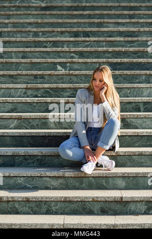 Young caucasian woman en contexte urbain. Fille blonde portant des vêtements décontractés dans la rue. Femme élégante avec veste et jeans bleu sitti Banque D'Images
