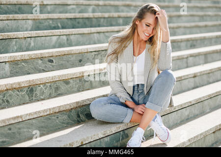 Beau young caucasian woman smiling in contexte urbain. Fille blonde portant des vêtements décontractés dans la rue. Femme élégante avec veste et jean bleu Banque D'Images