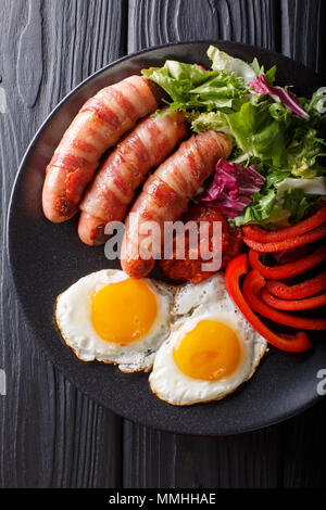 Les porcs en petit-déjeuner saucisses frites couvertures enrobé de bacon, d'œufs, de la sauce et salade fraîche gros plan sur une assiette. Haut Vertical Vue de dessus Banque D'Images