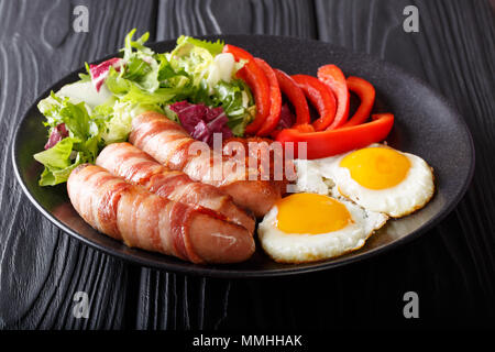 Petit-déjeuner anglais : Porcs dans des couvertures saucisses frites enrobé de bacon, oeufs, salade de légumes et sauce close-up sur une plaque sur une table horizontale. Banque D'Images