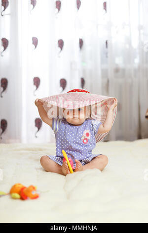 Un peu drôle portrait petite fille un enfant est assis à la maison sur le sol sur un tapis de lumière. Sur sa tête est un grand chapeau de paille d'été avec le soleil Banque D'Images