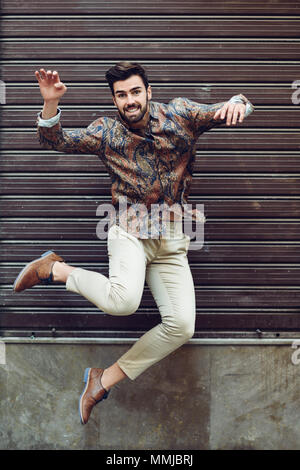 Jeune homme barbu qui saute dans l'arrière-plan urbain à bras ouverts portant des vêtements décontractés. Guy avec barbe et coiffure moderne dans la rue. Banque D'Images