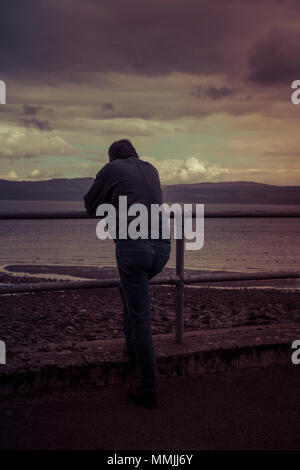 Silhouette of man leaning on railing surplombant la mer, avec des nuages sur les montagnes de l'autre côté de la baie, au crépuscule ou à l'aube Banque D'Images