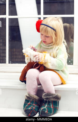 Petite fille dans un chandail tricoté avec une fleur et un bandage sur sa tête avec un jouet dans la main assis sur la fenêtre Banque D'Images