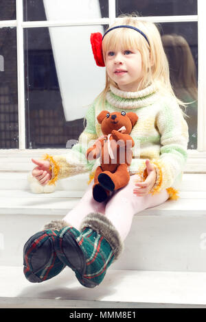 Petite fille blonde pullover en tricot dans une fleur et un bandage sur sa tête avec un jouet dans la main assis sur la fenêtre Banque D'Images