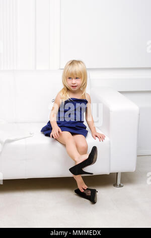 Petite fille de 3 ans assise sur un canapé blanc dans la robe de sa mère et des chaussures à talons hauts