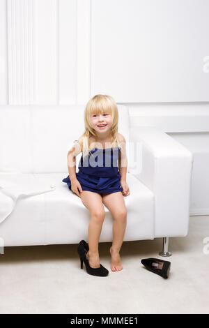 Petite fille de 3 ans assise sur un canapé blanc dans la robe de sa mère et des chaussures à talons hauts