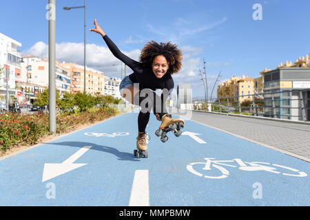 Jeune femme noire monter à cheval sur patins à l'extérieur, sur la ligne de vélo. Smiling girl avec coiffure afro à roues alignées sur un jour ensoleillé Banque D'Images