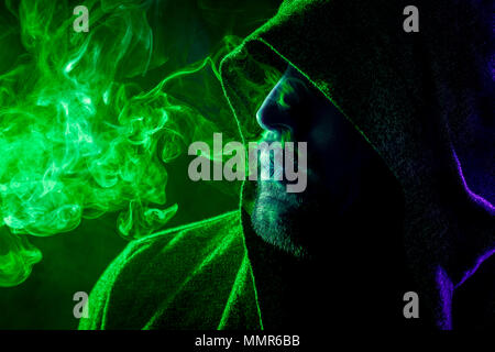Un bel homme dans un capot noir fume et exhale de gros nuages de fumée de couleur vert et bleu sur un fond isolé noir Banque D'Images