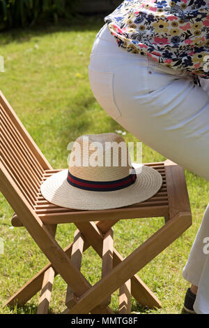 Femme sur le point de s'asseoir sur un chapeau de paille placé sur une chaise en bois dans un jardin de campagne. Banque D'Images