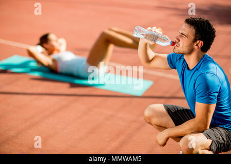 Jeune homme sportif de l'eau potable et de jeune femme faisant de l'exercice dans l'arrière-plan Banque D'Images