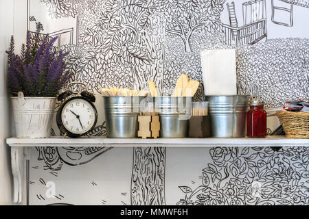 Plateau blanc avec des fleurs dans un seau, avec réveil et divers accessoires de cuisine sur mur blanc fond avec motif. Banque D'Images