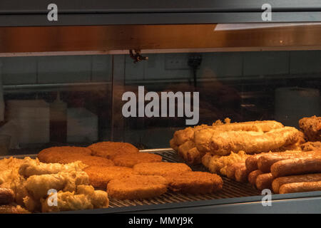 Les aliments frits dans une armoire chauffée dans un poisson et chip shop ou restauration rapide vente à emporter sur la high street. Les aliments frits hamburgers et des croquettes de poisson au chaud. Banque D'Images