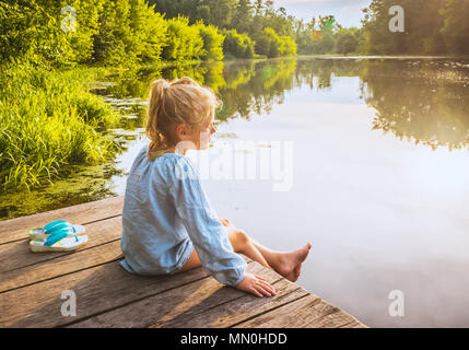 Petite fille solitaire rêvant sur un quai près de petit lac sur chaude journée d'été Banque D'Images