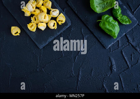 Maison fraîchement préparés flouring raw tortellini prêt à cuire sur la plaque de pierre sombre avec des feuilles de basilic sur fond noir. La cuisine traditionnelle italienne Banque D'Images