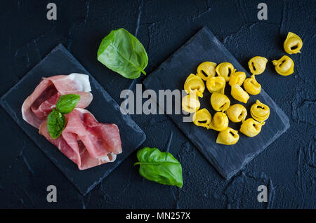 Matières premières de viande faits maison fraîchement préparés tortellini prosciutto rempli prêt à cuire sur la plaque de pierre sombre avec des feuilles de basilic sur fond noir. J'ai traditionnelle Banque D'Images