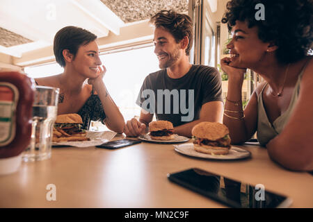 Groupe d'amis assis dans un restaurant avec un hamburger empilés sur la table. Heureux jeune homme avec ses amis féminins assis ensemble à café et hav Banque D'Images