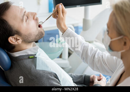 Portrait de femme dentiste dents l'examen à l'aide d'un miroir, se concentrer sur l'homme mûr assis dans fauteuil dentaire avec bouche ouverte Banque D'Images