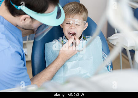 High angle portrait de petit garçon grimaçant de douleur assis dans fauteuil dentaire dentiste dents alors que son traitement et le remplissage de cavité clinique moderne Banque D'Images