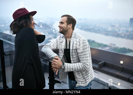 Joli couple interracial sur le balcon sur l'arrière-plan du paysage urbain. Black Girl in un crimson hat et son petit ami blanc à l'un l'autre Banque D'Images