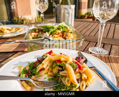 Table extérieure sous le soleil avec de la salade de pâtes servie sur assiette blanche, verres à vin en cristal et une bouteille de vin blanc frais dans le refroidisseur Banque D'Images