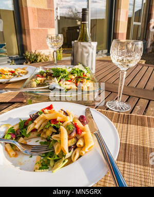 Table extérieure sous le soleil avec de la salade de pâtes servie sur assiette blanche, verres à vin en cristal et une bouteille de vin blanc frais dans le refroidisseur Banque D'Images