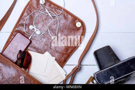 Sac en cuir marron femelle ouverte, un smartphone blanc et le casque est tombé, lunettes sur un fond de bois blanc Banque D'Images