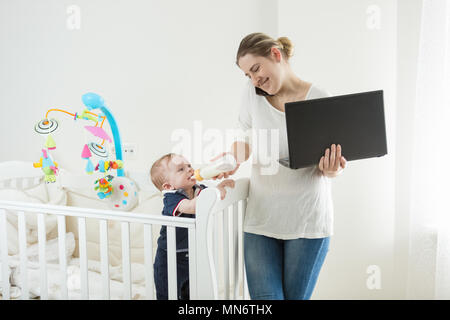 Portrait de jeune femme d'occupé à travailler sur ordinateur, de parler par téléphone et nourrir son bébé Banque D'Images