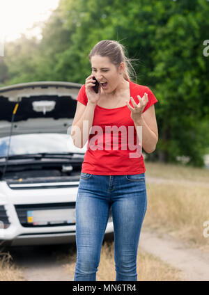 Angry young woman standing at voiture cassée dans le champ et de crier au téléphone mobile Banque D'Images