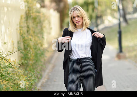 Belle blonde femme en milieu urbain. Jeune fille vêtu de noir veste blazer et pantalon à rayures debout dans la rue. Jolie femme avec les cheveux droits hairstyle et les yeux bleus. Banque D'Images