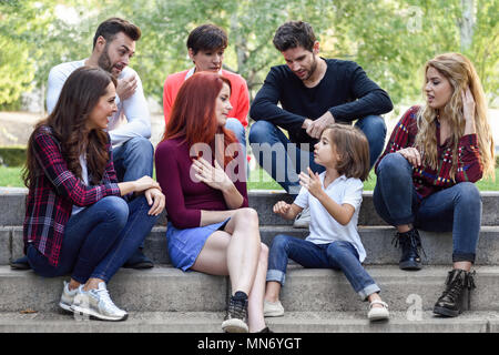 Les jeunes avec une petite fille de parler à l'extérieur en milieu urbain. Les femmes et les hommes assis dans les escaliers dans la rue portant des vêtements décontractés. Banque D'Images