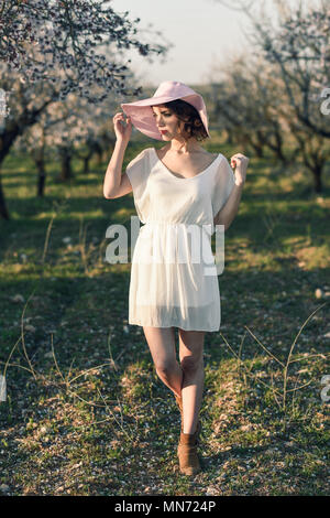Portrait de jeune femme dans le champ fleuri au printemps. Les fleurs d'amandiers en fleurs. Girl wearing robe blanche et chapeau de soleil rose Banque D'Images