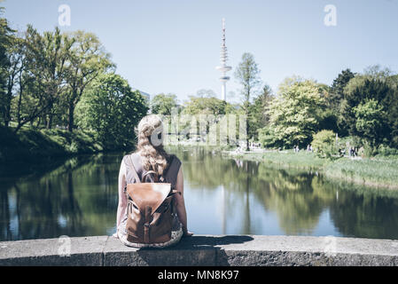Vue arrière de blonde woman sitting on wall par étang dans le parc aux beaux jours Banque D'Images