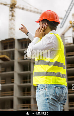 Jeune ingénieur en construction dans un casque de parler par téléphone et en pointant à l'édifice en construction Banque D'Images