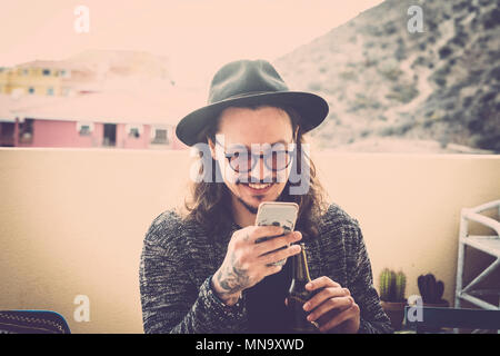 Longs cheveux beau jeune homme lire et contrôler le téléphone sur la terrasse en plein air. roofteop black hat et les lunettes et tatouage sur la main. fashion model nic sith Banque D'Images