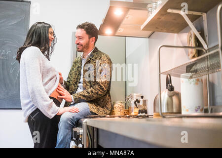 Femme enceinte de son mari l'homme assis sur la table de cuisine de toucher son corps et sourire et murmure à son amour ce qui concerne l'intérieur concept.. Banque D'Images