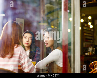 Trois professionnels de belles jeunes femmes asiatiques assis à table à discuter en conversation - café ou thé, tourné à travers une vitre. Banque D'Images