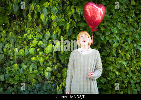 Jeune femme ballon en forme de coeur debout contre des plantes Banque D'Images