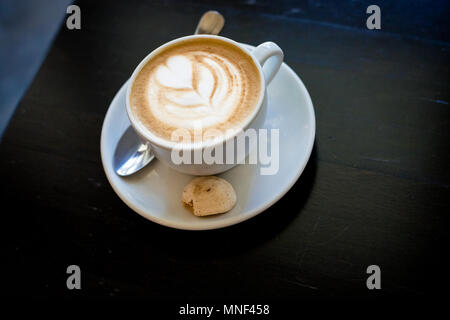 Cappuchino ou café latte dans une tasse en forme de coeur blanc avec mousse et cookies,verre du matin sur table en bois.Le café dans un mug blanc sur fond sombre Banque D'Images