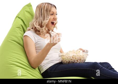 Joyful jeune femme assise sur un pouf poire et eating popcorn isolé sur fond blanc Banque D'Images