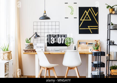 Or noir et blanc suspendu au-dessus de l'affiche de la géométrique et espace bureau en bois dans un style scandinave avec des chaises de créateur, plante monstera et industrielle Banque D'Images